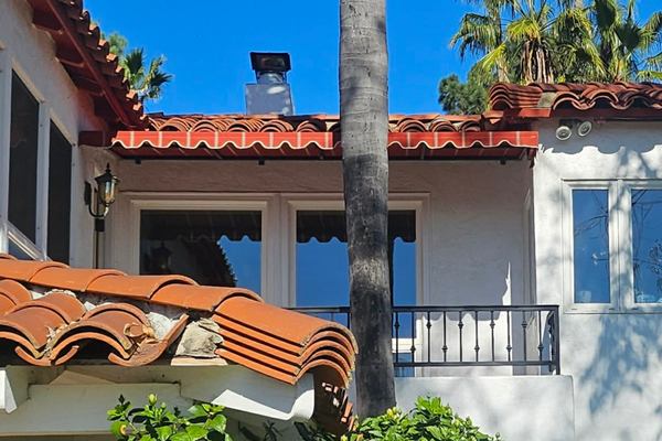 A Spanish-style house with red tiles and white walls has a striped awning over a balcony. A palm tree stands in front against a blue sky.